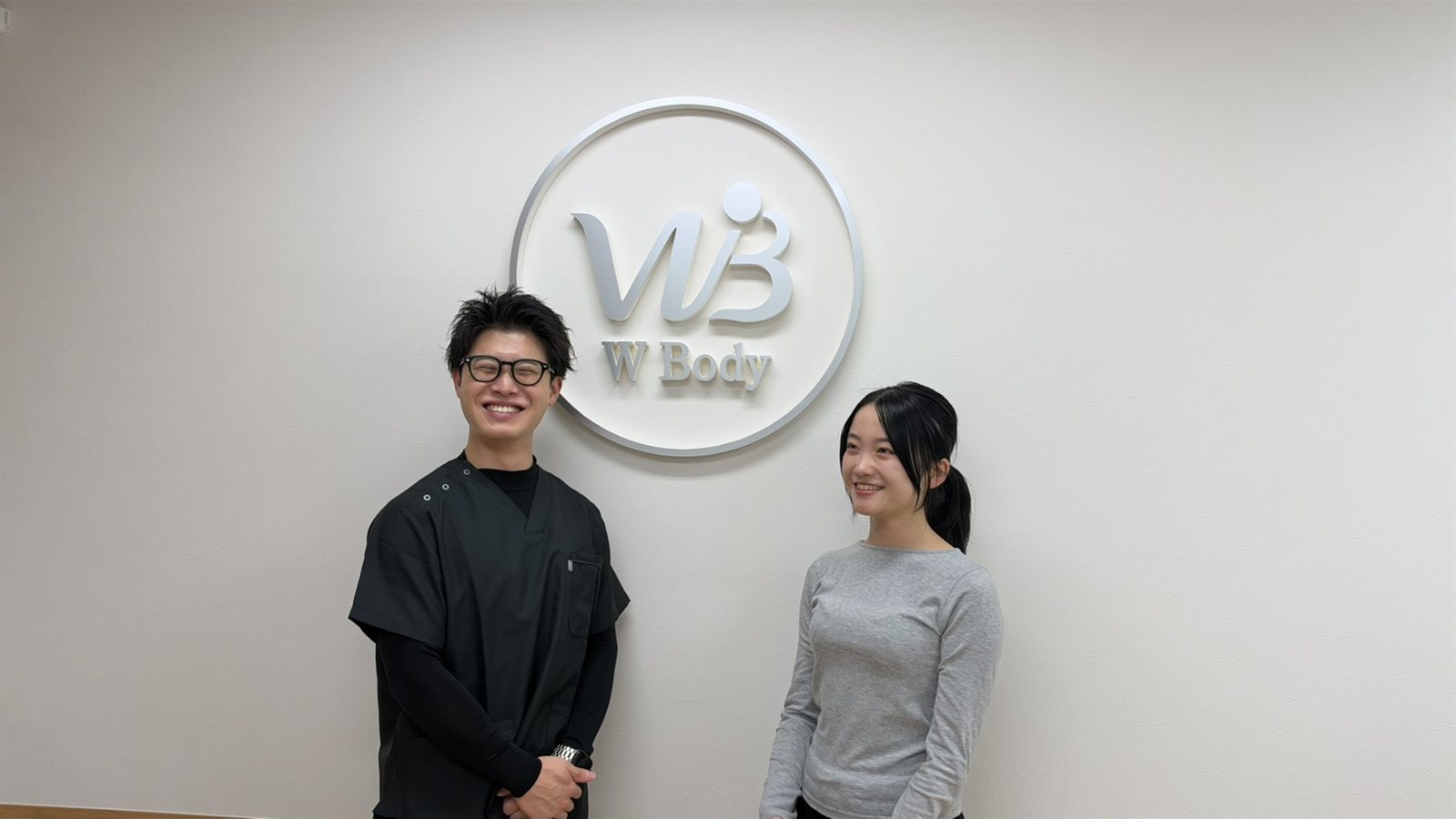 W Body のロゴの前で笑顔で話す理学療法士とお客様